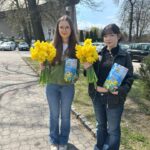 Two young women stand on a sunny sidewalk outdoors, each holding a bouquet of bright yellow flowers and a blue brochure.