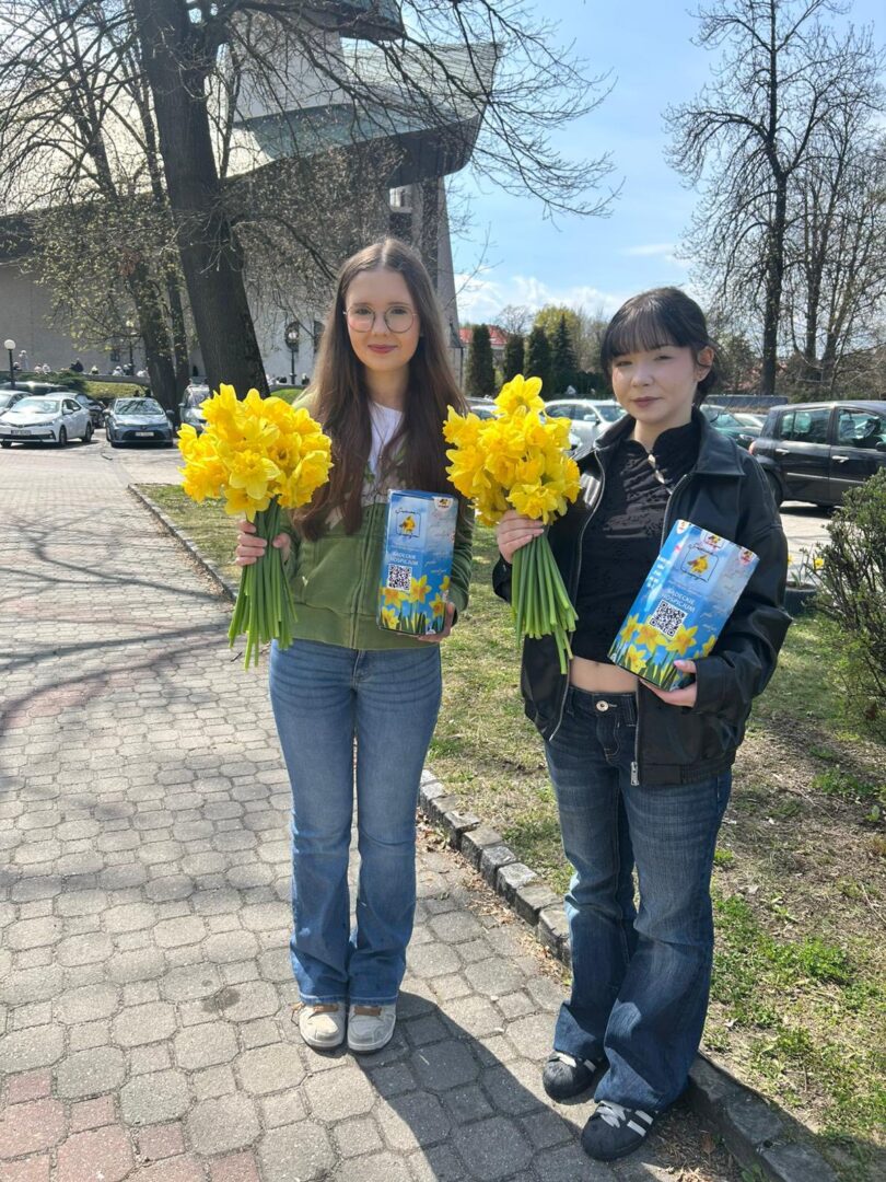 Two young women stand on a sunny sidewalk outdoors, each holding a bouquet of bright yellow flowers and a blue brochure.