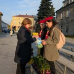 Older woman hands yellow flowers to two young volunteers in red berets at an outdoor town square; potted plants on a bench nearby.