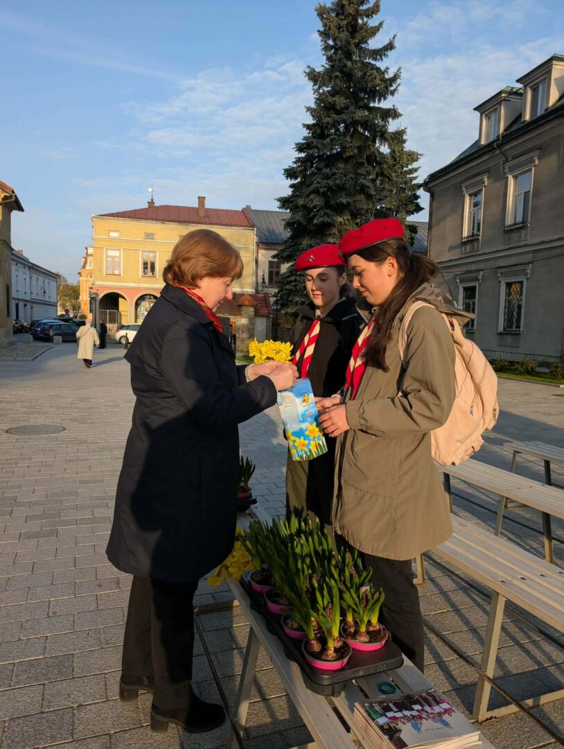 Older woman hands yellow flowers to two young volunteers in red berets at an outdoor town square; potted plants on a bench nearby.