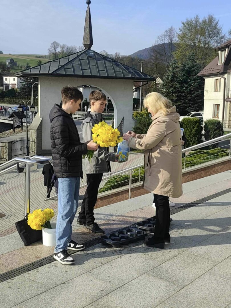 Two boys handing a bouquet of bright yellow flowers to a woman on a sunny outdoor walkway.
