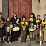 Eight children stand in a row outside a building with large wooden doors, each holding yellow daffodil bouquets and wicker baskets.