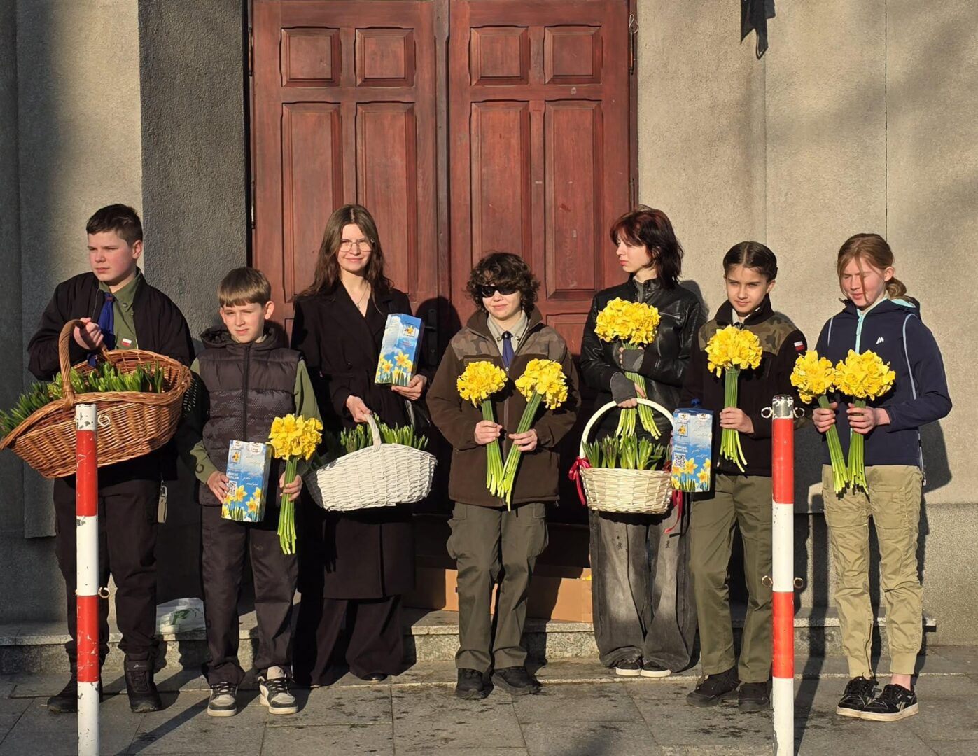 Eight children stand in a row in front of a wooden door, each holding bright yellow daffodils and gift baskets outdoors.