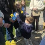 Child hands a yellow daffodil to a taller woman while others watch outdoors on a sunny day.