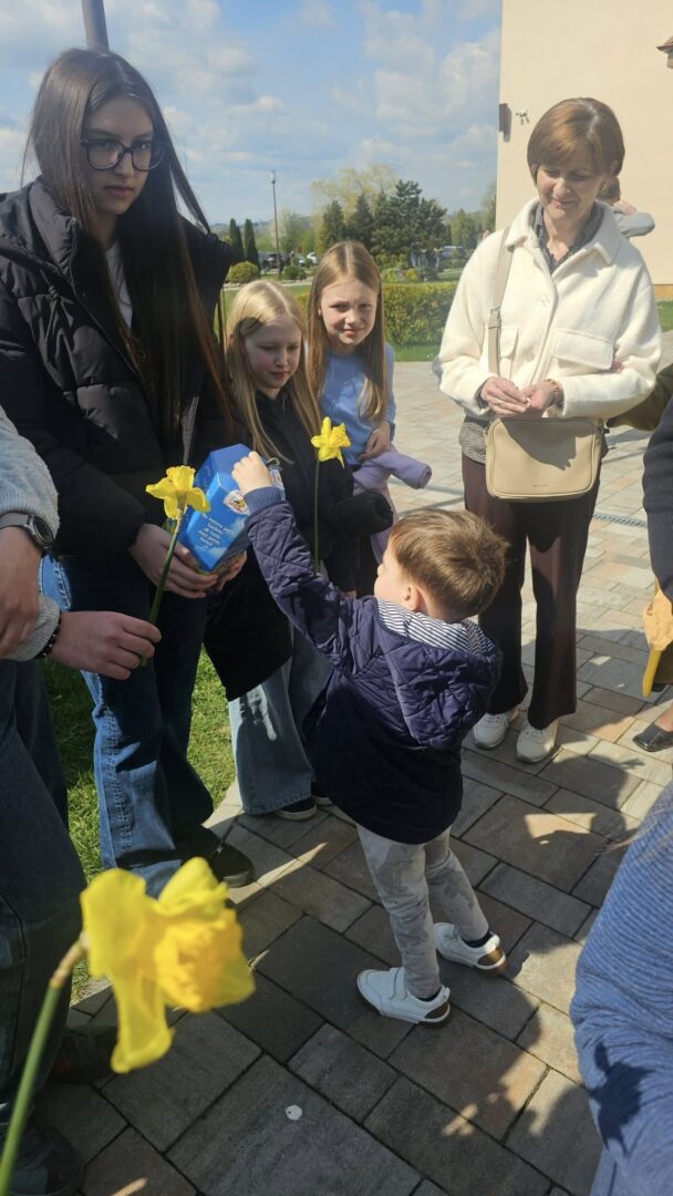 Child hands a yellow daffodil to a taller woman while others watch outdoors on a sunny day.