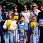 Eight people posing outdoors in bright daylight; three children in front hold yellow daffodils and blue gift boxes, with four adults behind them in casual to smart-casual clothing.