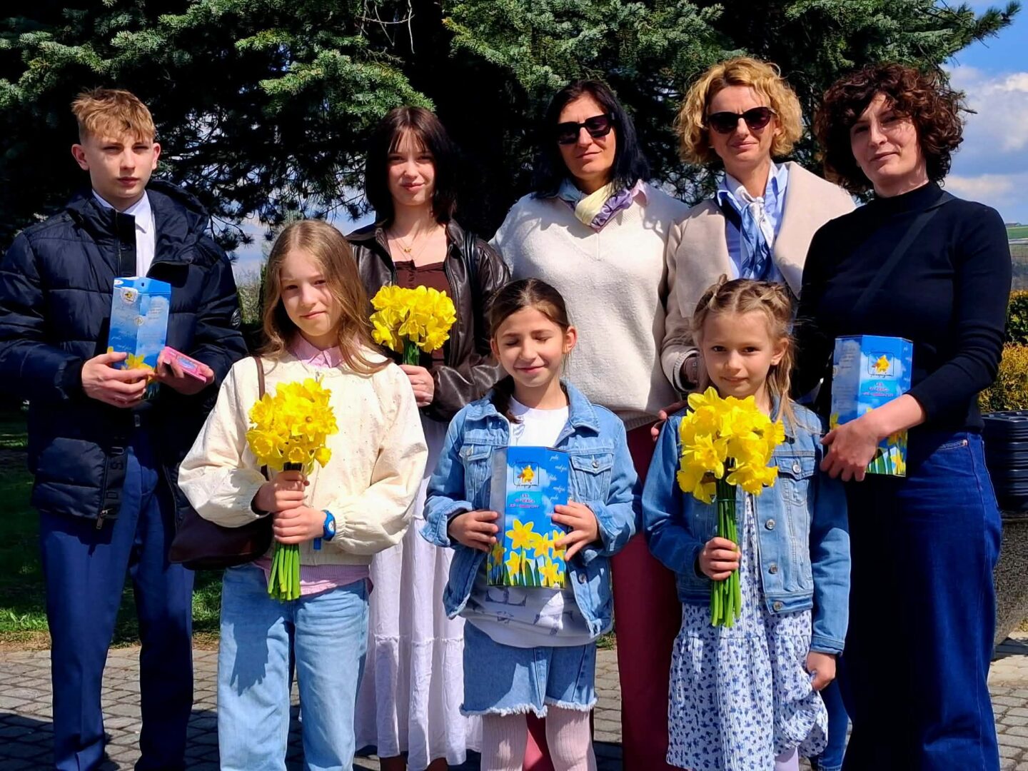 Eight people posing outdoors in bright daylight; three children in front hold yellow daffodils and blue gift boxes, with four adults behind them in casual to smart-casual clothing.