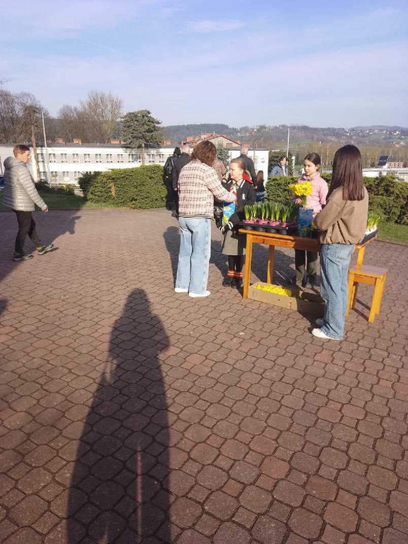 Group of people gathered around a wooden table displaying potted flowers on a sunny brick-paved outdoor area.