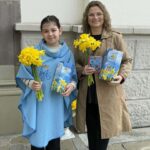 Two people stand outside a building, each holding a bouquet of yellow daffodils and a blue booklet for a spring event.