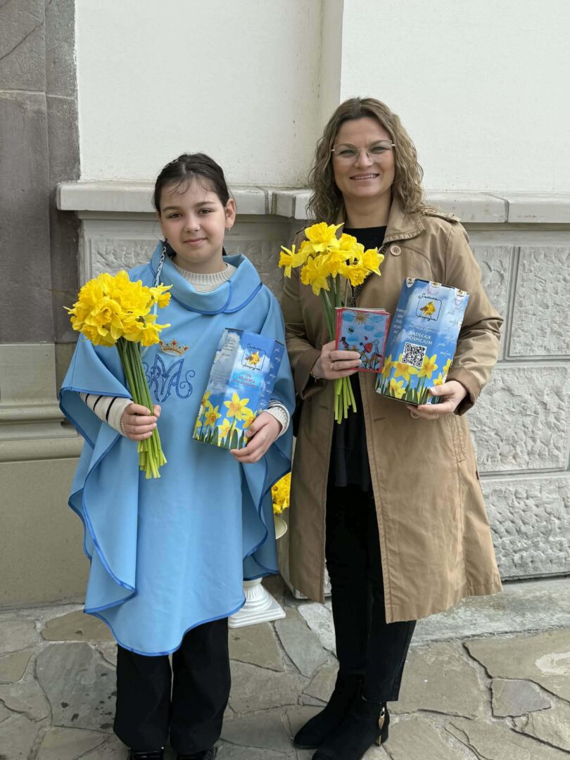 Two people stand outside a building, each holding a bouquet of yellow daffodils and a blue booklet for a spring event.