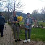 Woman in a checkered suit holding a bouquet of yellow flowers stands on a brick path in a park, with a boy in a gray suit beside her.