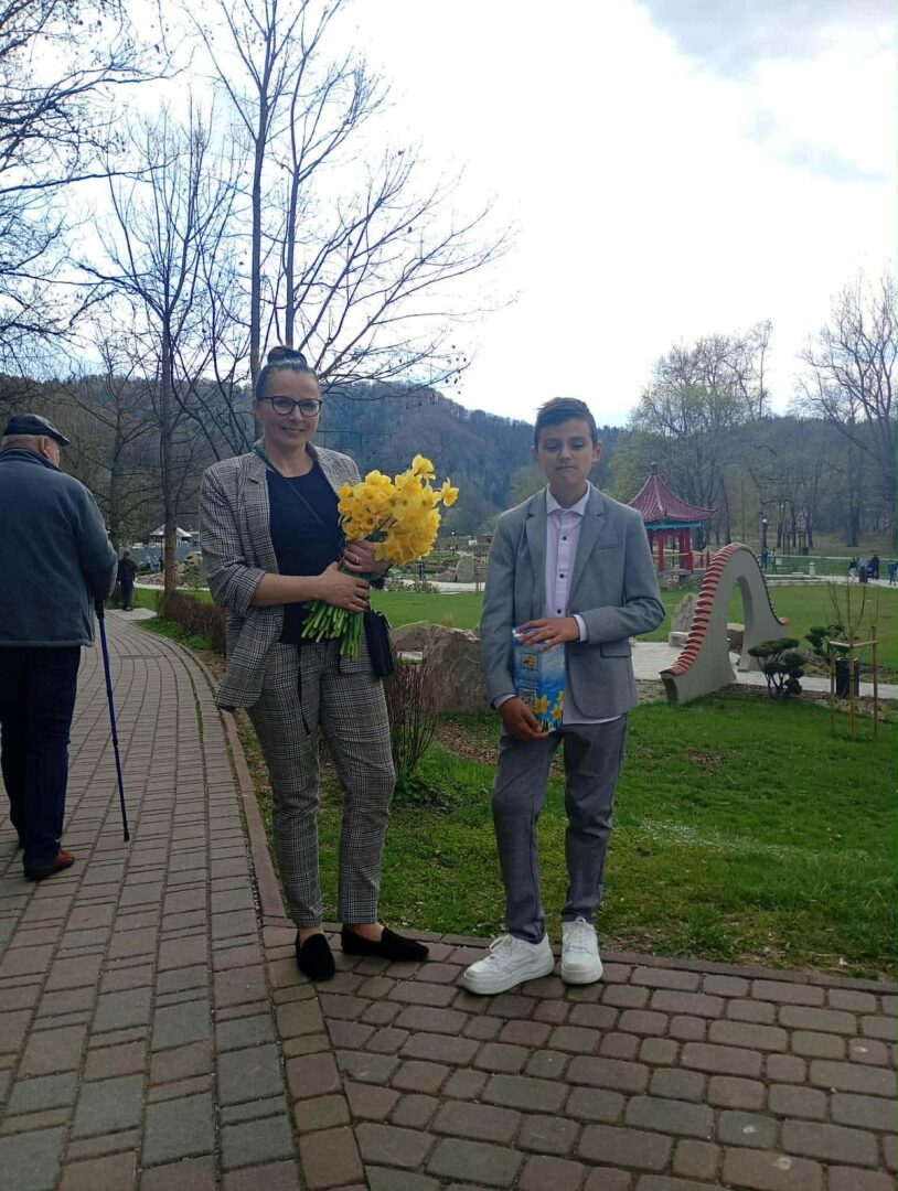 Woman in a checkered suit holding a bouquet of yellow flowers stands on a brick path in a park, with a boy in a gray suit beside her.