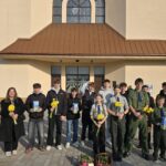 Group of students standing outside a church or school building, each holding yellow daffodils. Sizeable crowd in casual and uniform outfits.