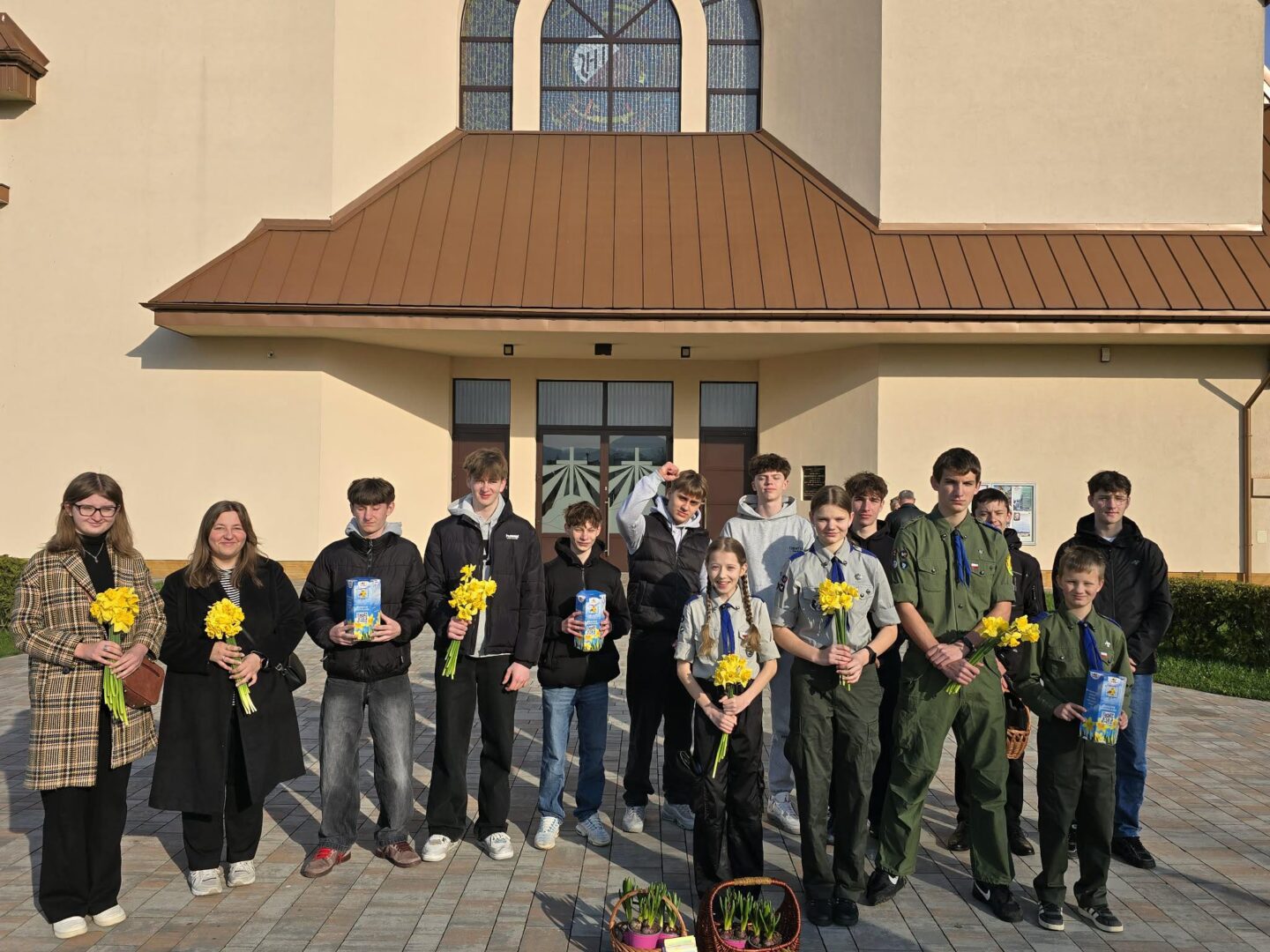Group of students posing outside a beige building with a brown, slanted roof, each holding a yellow daffodil bouquet.