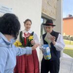 Two people in traditional Polish folk costumes greet a passerby outside a museum, with a man holding a brochure and a blue box while a woman holds yellow flowers.