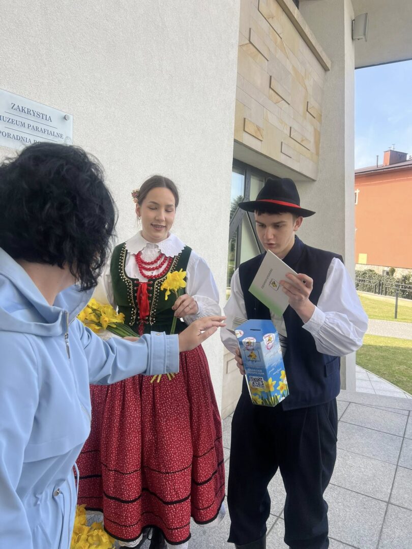 Two people in traditional Polish folk costumes greet a passerby outside a museum, with a man holding a brochure and a blue box while a woman holds yellow flowers.