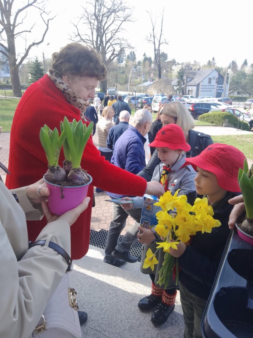 Woman in a red coat hands yellow daffodils to children wearing red hats at an outdoor event.
