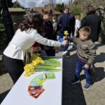 Woman in white hands a yellow daffodil to a boy at an outdoor table; others wait nearby.