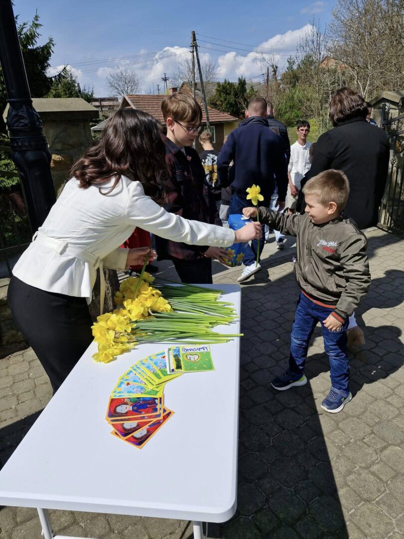 Woman in white hands a yellow daffodil to a boy at an outdoor table; others wait nearby.