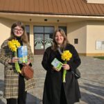 Two women stand outside a beige building holding yellow daffodils and books, smiling at the camera.