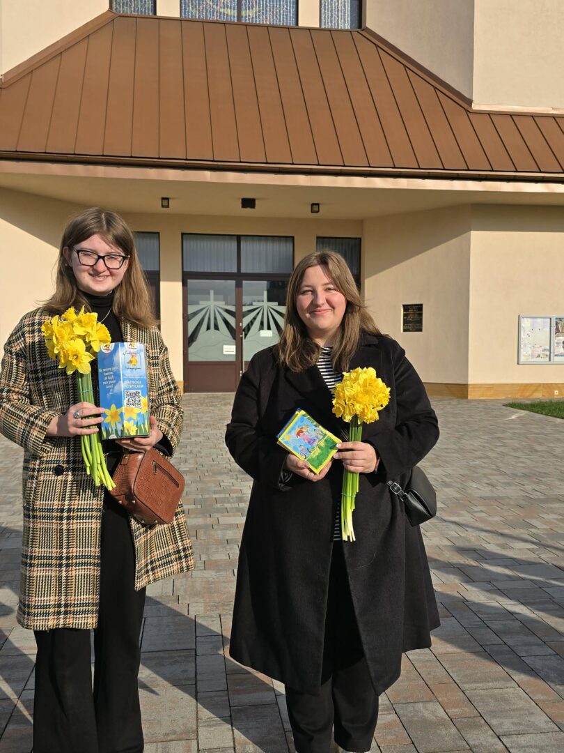 Two women stand outside a beige building holding yellow daffodils and books, smiling at the camera.