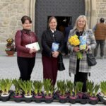 Three women stand outside a stone church doorway behind a table with potted plants; one holds yellow daffodils and a book.