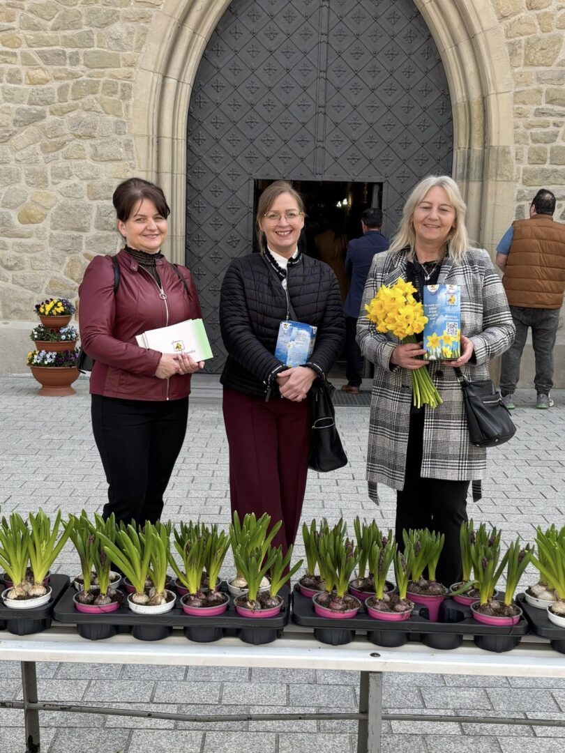 Three women stand outside a stone church doorway behind a table with potted plants; one holds yellow daffodils and a book.