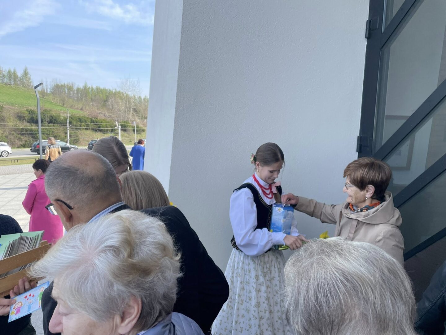 Girl in a traditional folk dress receives a gift from an older woman outside a building, with a small crowd nearby.