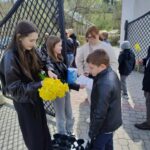 A girl in a black jacket hands yellow daffodils to a boy in a leather jacket outdoors by a gate.