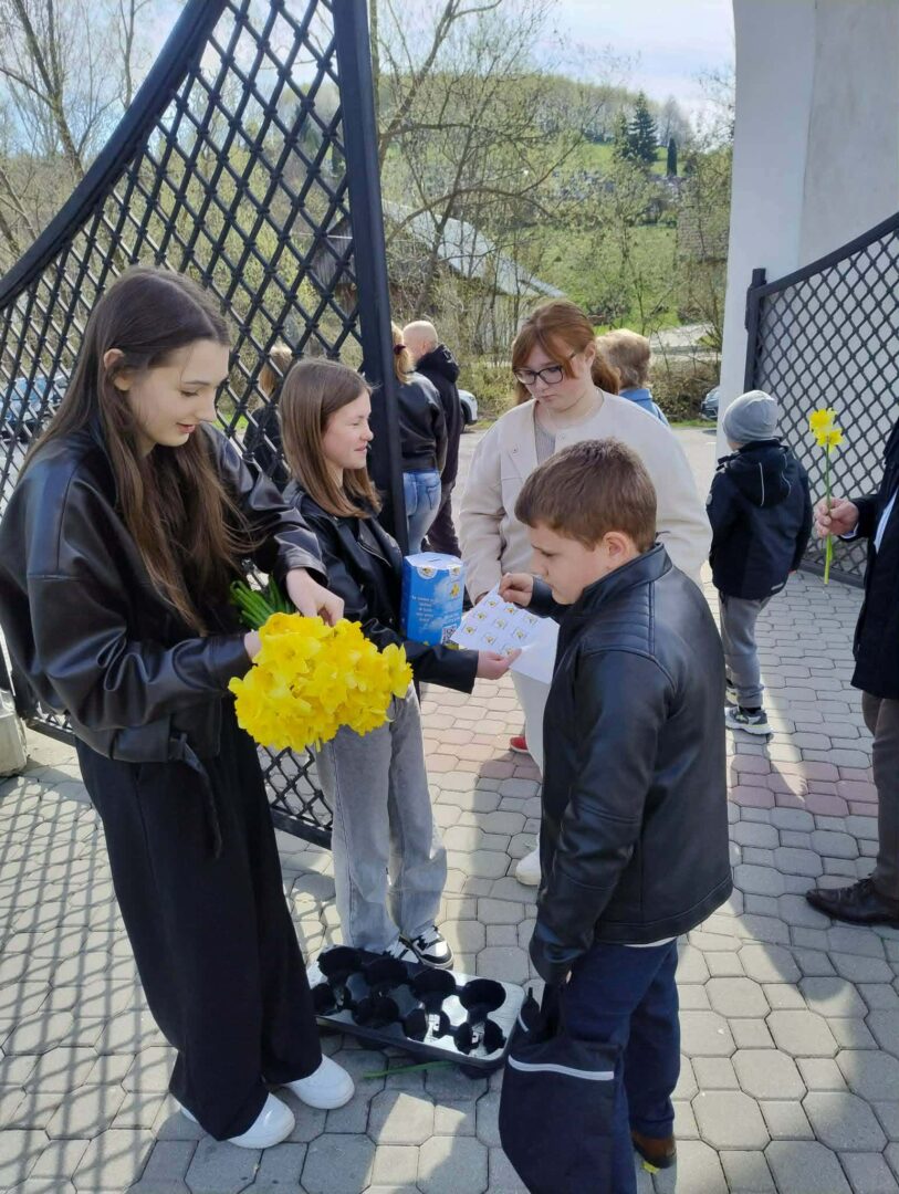 A girl in a black jacket hands yellow daffodils to a boy in a leather jacket outdoors by a gate.