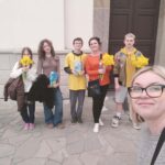Woman with glasses taking a selfie with five kids outside a stone building; each child holds yellow flowers.