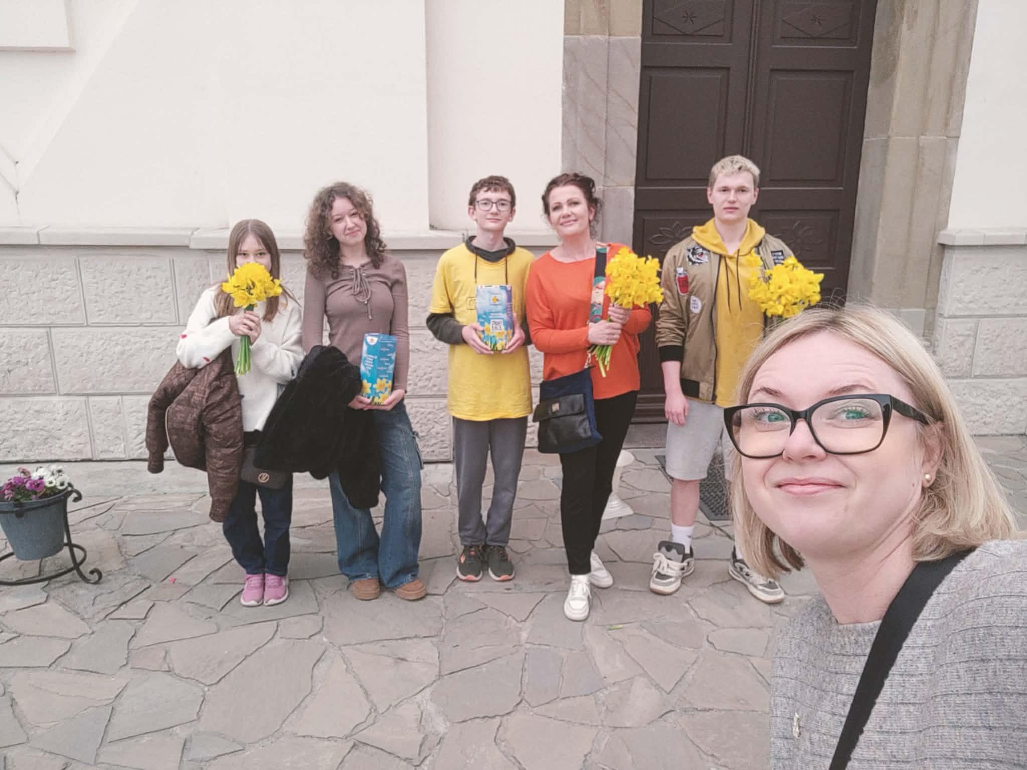 Woman with glasses taking a selfie with five kids outside a stone building; each child holds yellow flowers.