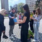 People at an outdoor community event in a sunny plaza; a man hands a blue item to a woman in a black outfit while others hold plants nearby, with a tall apartment building in the background.
