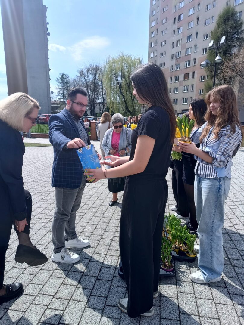 People at an outdoor community event in a sunny plaza; a man hands a blue item to a woman in a black outfit while others hold plants nearby, with a tall apartment building in the background.
