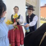 Woman in a green folk vest and red skirt hands yellow daffodils to a visitor while a man in a black hat offers a card, outside a modern building.