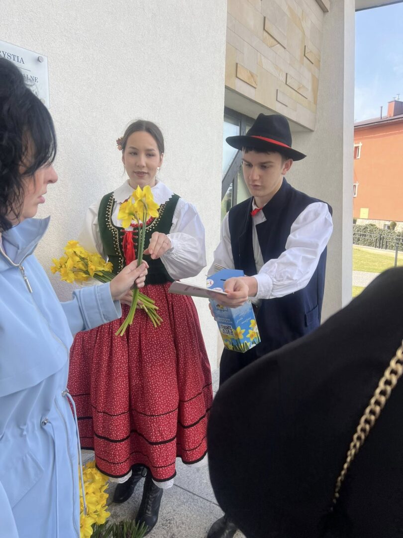 Woman in a green folk vest and red skirt hands yellow daffodils to a visitor while a man in a black hat offers a card, outside a modern building.