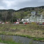Line of red fire trucks parked along a rural road as smoke rises from houses in a hillside village.