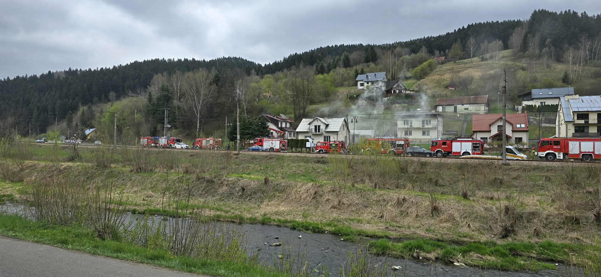 Line of red fire trucks parked along a rural road as smoke rises from houses in a hillside village.