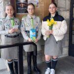 Three girls in scout uniforms pose outside a stone wall bulletin board; one holds a card, another a blue gift box, and the third holds a bouquet of yellow daffodils.