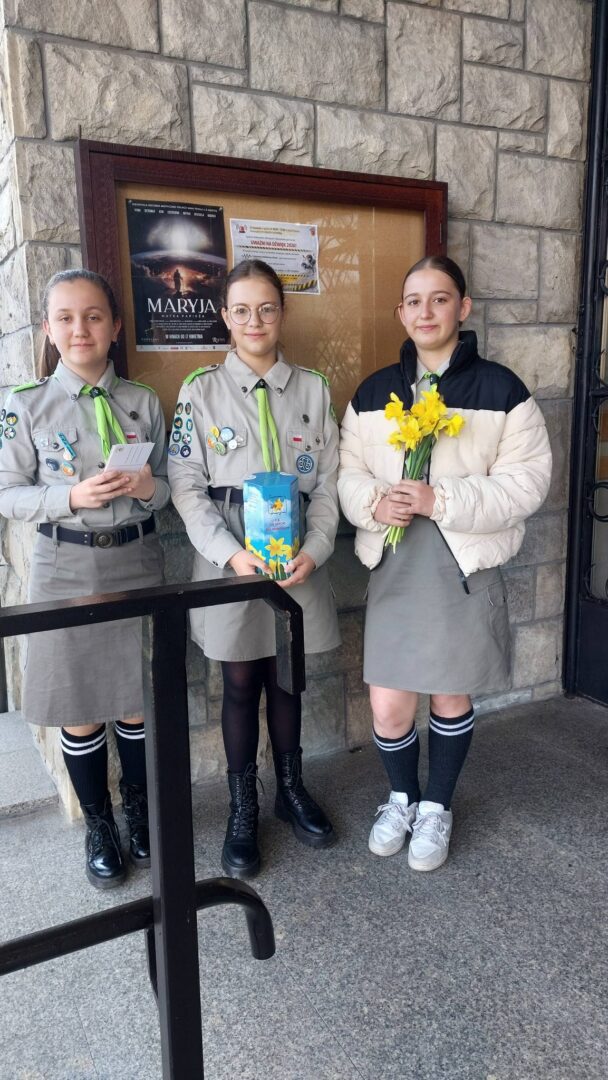 Three girls in scout uniforms pose outside a stone wall bulletin board; one holds a card, another a blue gift box, and the third holds a bouquet of yellow daffodils.