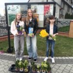 Three kids stand outside by a community notice board, each holding flowers: blue hyacinths in a purple pot, yellow tulips, and a bouquet of yellow daffodils.