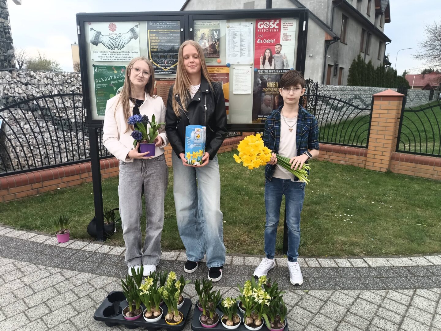 Three kids stand outside by a community notice board, each holding flowers: blue hyacinths in a purple pot, yellow tulips, and a bouquet of yellow daffodils.