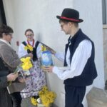 People in traditional dress share flowers and gifts outside a building; a man in a dark vest and hat offers a package to an older woman and a younger woman in folk attire holds a yellow daffodil bouquet.
