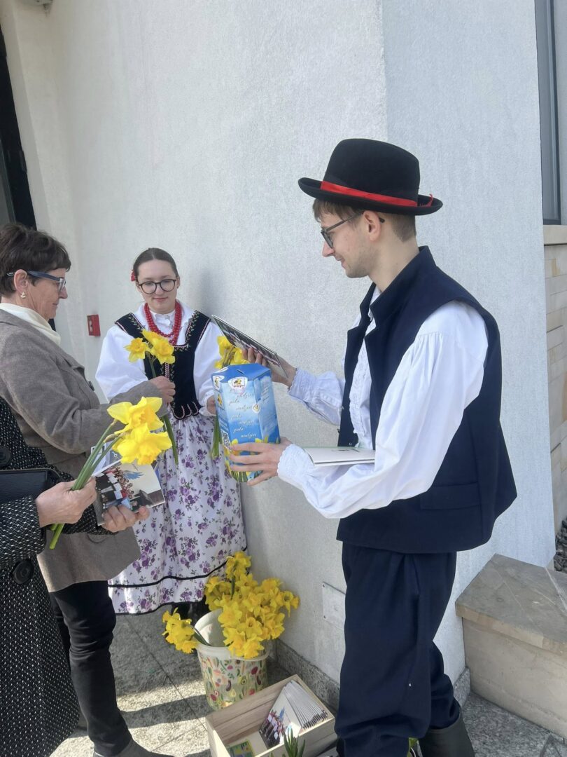 People in traditional dress share flowers and gifts outside a building; a man in a dark vest and hat offers a package to an older woman and a younger woman in folk attire holds a yellow daffodil bouquet.