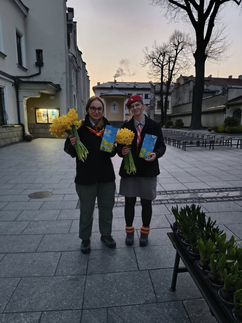 Two people in scout-like uniforms stand in a paved outdoor plaza at dusk, each holding a bouquet of yellow daffodils and a blue pamphlet.