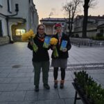 Two people in uniforms stand in a paved town square at dusk, each holding a bouquet of yellow daffodils and a blue flyer/pamphlet.