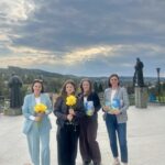Four women stand on a spacious outdoor terrace, two holding yellow flowers and two holding books, with a red bucket of flowers nearby and statues in the background under a cloudy sky.