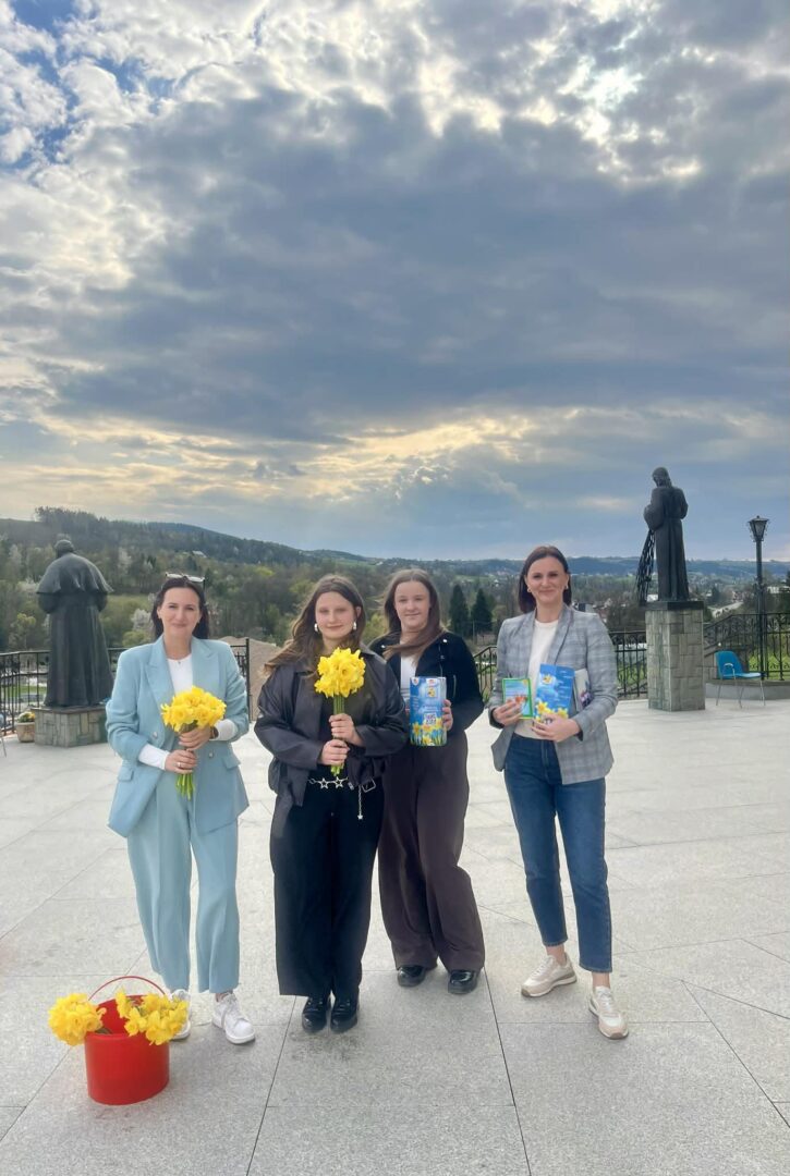 Four women stand on a spacious outdoor terrace, two holding yellow flowers and two holding books, with a red bucket of flowers nearby and statues in the background under a cloudy sky.