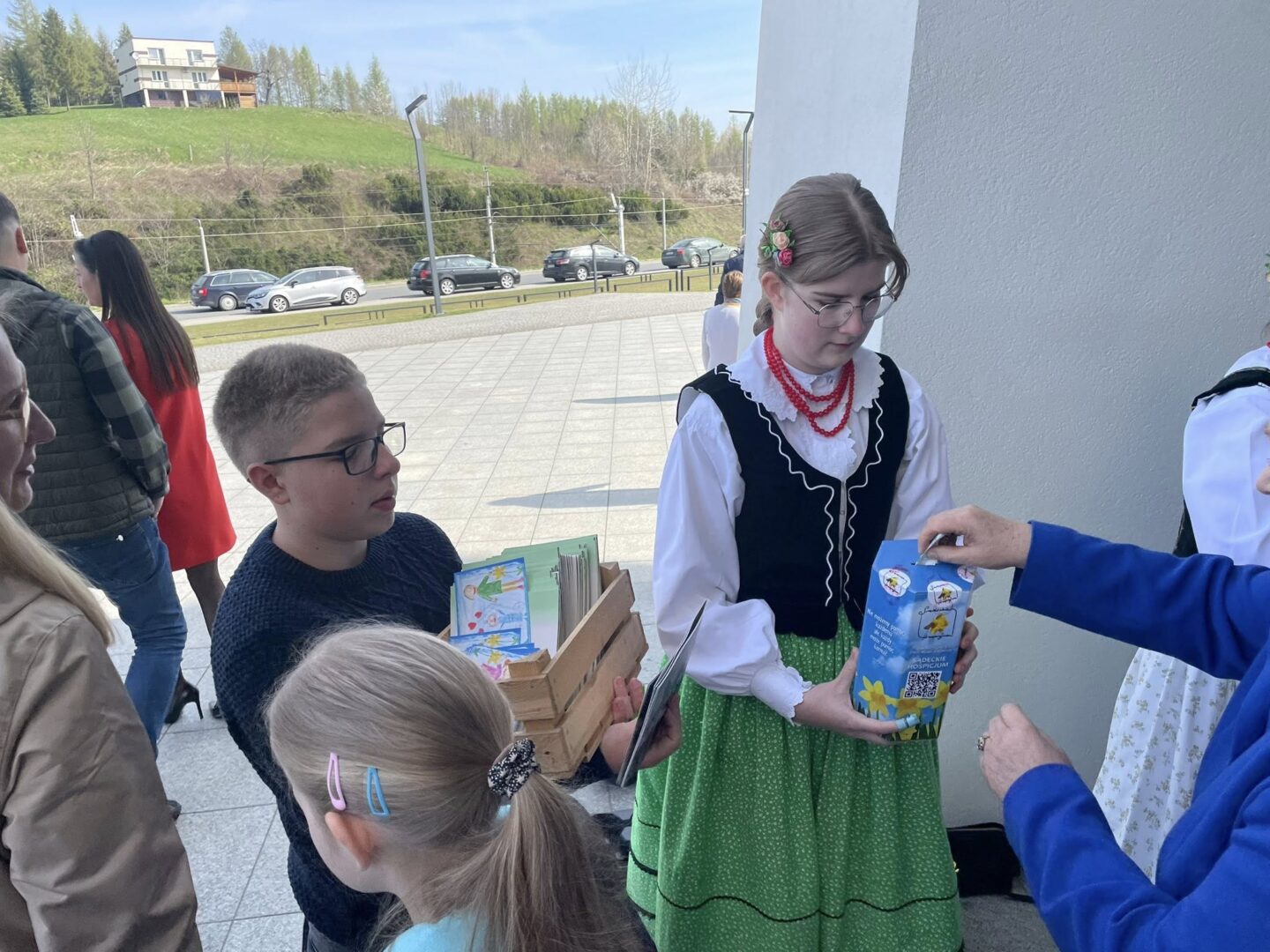 Girl in a traditional green- and white-outfit with red bead necklace hands a carton to a person outside a building while others wait nearby.