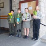 Four children in scout uniforms stand on stone steps outside a building, holding flowers and a card.